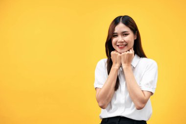 Beyaz kısa kollu gömlek giyen genç Asyalı kadın ve iki eliyle çenesine dokunup gülümserken sarı arka planda tek başına duruyor..