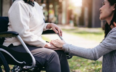 Tekerlekli sandalyedeki son sınıf öğrencisi ve aile kavramı, genç Asyalı kadın, anneyi tekerlekli sandalyede cesaretlendirmek için el ele tutuşurken, yaşlı kadın ve kız bahçede konuşurken dinleniyor..