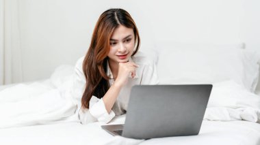 Ev eğlencesi yaşam tarzı konsepti, filmi günlük kıyafetlerle izleyen ve evinde rahat bir yaşam tarzıyla rahat bir yatakta uzanırken bilgisayarında arkadaşlarıyla sohbet eden genç bir kadın..