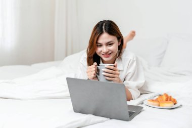 Evde eğlence yaşam tarzı konsepti. Sıcak çikolata içen, dizüstü bilgisayarda film izleyen ve evde rahat bir yaşam tarzıyla rahat bir şekilde uzanan genç bir kadın..