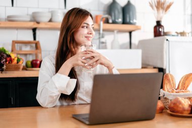 Ev eğlencesi yaşam tarzı konsepti, günlük giysiler içinde mutfak odasında oturan süt içen ve dizüstü bilgisayarla film seyreden evde yaşam tarzı olan genç bir kadın..