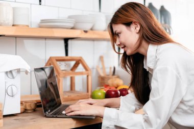 Ev eğlencesi yaşam tarzı konsepti. Mutfak odasında sıradan giyinmiş genç bir kadın. Bilgisayarda arkadaşlarıyla yazışıyor ve sohbet ediyor..