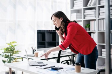 Akıllı telefon kullanan genç iş kadını strateji analizi ve ofiste yeni iş planları yaparken bilgisayardan iş verilerine bakıyor..