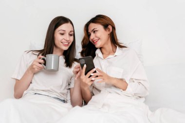 Genç Asyalı çift internette sörf yapmak için akıllı telefon kullanıyor ve battaniyenin altında yatarken sıcak çikolata içiyor ve yatak odasındaki rahat yastıklara yaslanıyor..