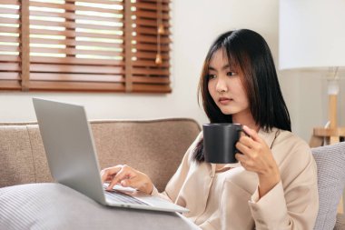 Kadın girişimci büyük rahat bir koltukta oturuyor ve evinde dinlenirken yastığa dizüstü bilgisayar koyup çalışmaya ve kahve içmeye başlıyor..