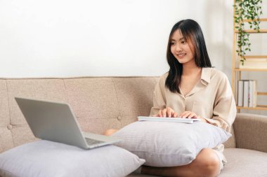 Kadın girişimci büyük rahat bir koltukta oturuyor ve evinde çalışırken iş verilerini daktilo etmek için dizüstü bilgisayarını ve klavyesini yastığa koyuyor..