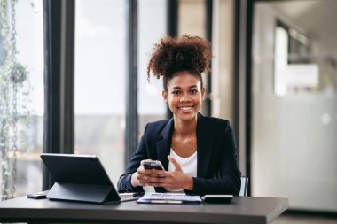 Takım elbiseli Afrikalı Amerikalı iş kadını akıllı telefondan muhasebe bilgilerini okuyor ve modern çalışma alanında evrak işleriyle uğraşırken projenin stratejisini düşünüyor..