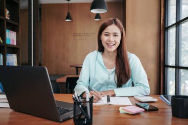Dişi öğrenci, dizüstü bilgisayarda öğrenim görürken ve sınav hazırlıkları için ödev yaparken kütüphanedeki not defterine not alıyor..