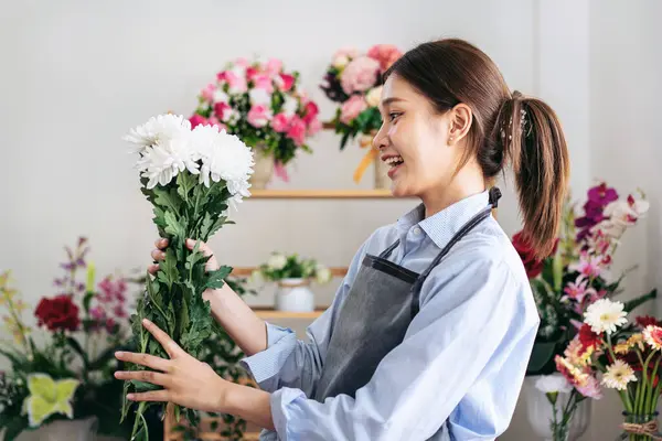 Female florist in apron holding white chrysanthemum with enjoying to creating and designing floral for arrangement flower bouquet on the table in her flower shop.