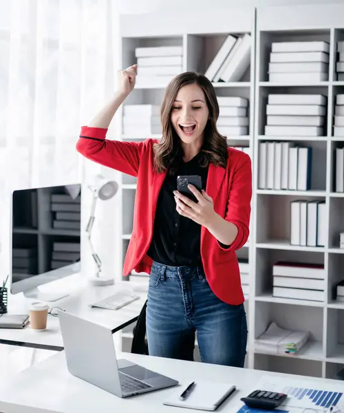 Accounting business concept, Accountant woman reads good news on smartphone with excited and raising fist to celebrate achievement while working about investment with business planning finance.