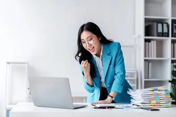 Accounting business concept, Accountant woman reads good news on laptop with excited and raising fist to celebrate achievement while working about investment with business planning finance.
