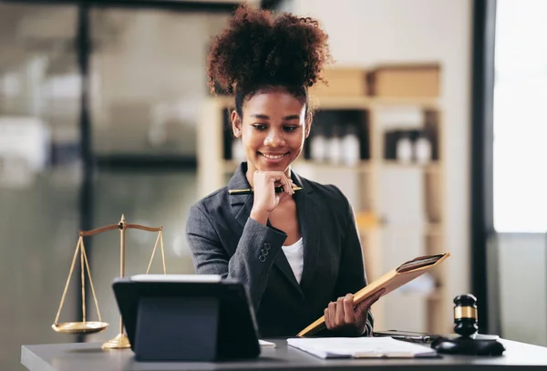 African american lawyer woman in suit holding envelope of business contract and reading information on tablet while sitting to working about legal and justice with judge hammer on desk in courtroom.