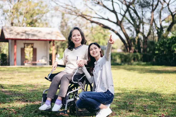 Senior on wheelchair and daughter family concept, Senior woman sitting on wheelchair to relaxation in the garden while mother and daughter pointing to watching nature view with enjoying.