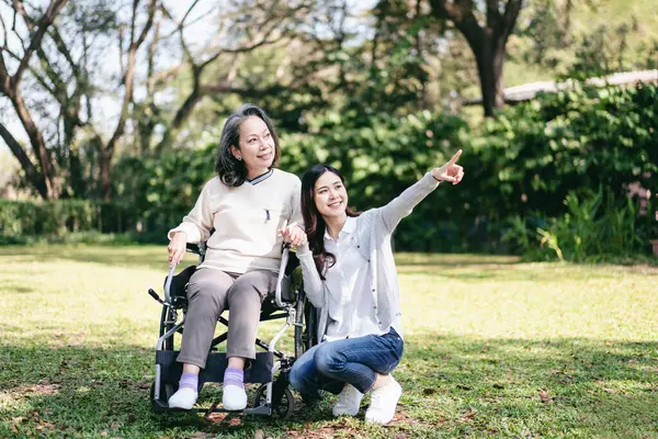Senior on wheelchair and daughter family concept, Senior woman sitting on wheelchair to relaxation in the garden while mother and daughter pointing to watching nature view with enjoying.