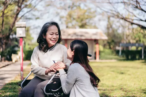 Senior on wheelchair and daughter family concept, Young asian woman holding hands to encourage mother on wheelchair while senior woman and daughter relaxation with talking in the park.