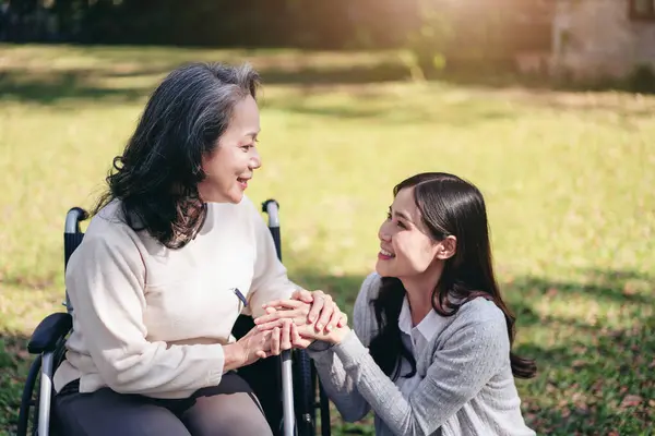 Senior on wheelchair and daughter family concept, Young asian woman holding hands to encourage mother on wheelchair while senior woman and daughter relaxation with talking in the park.