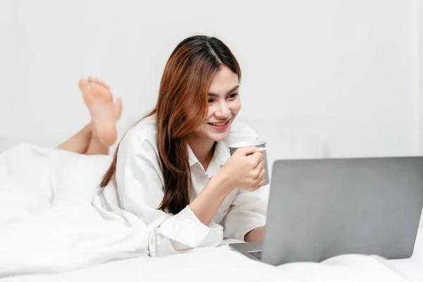 Activity at home leisure lifestyle concept, Young woman in clothes casual drinking hot chocolate and watching the movie on laptop while lying on comfy bed to leisure with lifestyle at home.