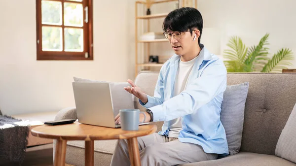 Businessman in casual is sitting comfortable sofa and wearing headphones to explaining business information with colleague while using laptop to meeting in video conference at home.