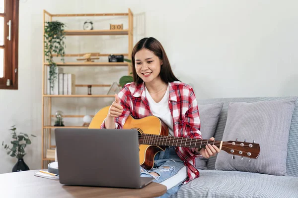 Young woman is playing guitar and making thumbs up with teachers after taking online guitar lessons on laptop while sitting on comfortable the couch in living room at home.