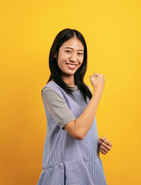 Sarı arka planda neşeli bir şekilde izole edilmiş genç Asyalı kadın gülümsüyor ve kutlama için yumruklarını kaldırıyor..
