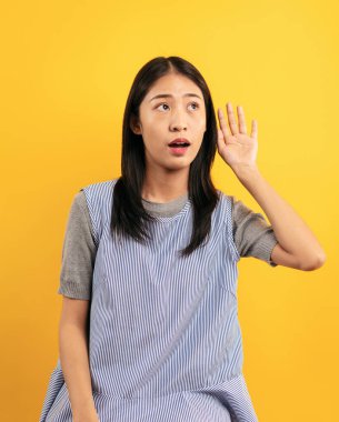 Sıradan kıyafetler içindeki genç Asyalı kadın dedikodu dinlerken ya da kulağına fısıldarken heyecanlı yüzüyle sarı arka planda izole edilmiş bir şekilde elini kulağına koyuyor..