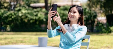 Bahçe konseptinde gevşeme yaşam tarzı, kulaklık takan Asyalı genç bir kadın ve arkadaşlarıyla video konferansında bahçede dinlenirken akıllı telefon kullanması..