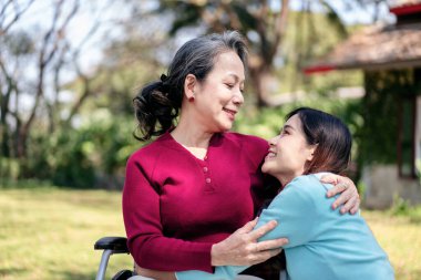 Tekerlekli sandalyedeki son sınıf öğrencisi ve aile konsepti. Genç Asyalı kadın bahçede annesiyle gülümsüyor ve ona sarılıyor..