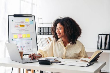 Masasında dizüstü bilgisayarı ve kitabı olan bir kadın oturuyor. Gülümsüyor ve işinden zevk alıyor. Masa bir hesap makinesi, bir fincan ve birkaç kitap da dahil olmak üzere çeşitli parçalarla dolu.