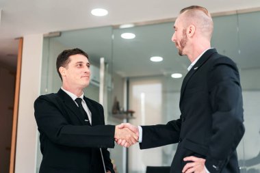 İş ortamında el sıkışan iki adam. Sahne profesyonel ve resmi. Tokalaşma, iki adam arasındaki saygı ve uzlaşmanın göstergesidir.