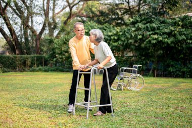 Bir adam ve bir kadın parkta yürüyorlar. Adam yürüteciyle kadına yardım ediyor.