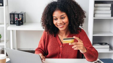 Bir kadın dizüstü bilgisayarın önünde kredi kartı tutarken gülümsüyor. Muhtemelen kartı internetten alışveriş yapmak için kullanıyor.