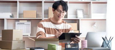 Erkek girişimci, evdeki alışveriş merkezinde çevrimiçi alışveriş yaparken müşteri göndermek için karton kutulara paketlemeden önce ürünleri kontrol etmek için bir pano tutuyor..