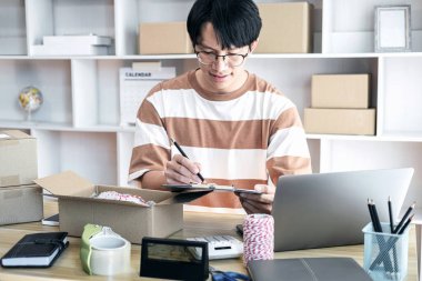 Erkek girişimci, evdeki alışveriş merkezinde çevrimiçi alışveriş yaparken müşteri göndermek için karton kutulara paketlemeden önce ürünleri kontrol etmek için bir pano tutuyor..