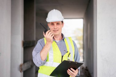 Sarı yelek ve şapka giyen bir adam elinde bir dosya ve bir telsiz tutuyor.