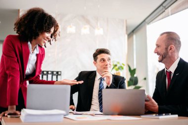 Üç adam ve bir kadın dizüstü bilgisayarları ve kağıtlarıyla bir masada oturuyorlar. Kadın gülümsüyor ve erkekler ona bakıyor. Sahne pozitif ve işbirlikçi.