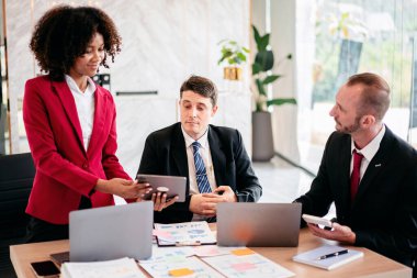 Üç erkek ve bir kadın dizüstü bilgisayar ve tabletlerle bir masada oturuyorlar. Kadın, erkeklerden birine bir tablet teslim ediyor. Sahne profesyonel ve odaklı.