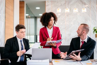 Takım elbiseli üç kişi önlerinde kırmızı ceketli bir kadınla bir masada oturuyorlar. Kadın elinde bir dosya ve kalem tutuyordu ve erkekler ona bakıyordu.
