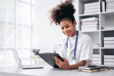 Beyaz laboratuvar önlüğü giyen bir kadın bir masada dizüstü bilgisayar ve tabletle oturuyor. Gülümsüyor ve işinden zevk alıyor.
