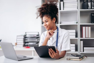 Beyaz laboratuvar önlüklü bir kadın bir masada dizüstü bilgisayar ve tabletle oturuyor. Dizüstü bilgisayar ekranına bakıyor ve şöyle düşünüyor:
