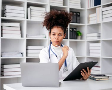 Bir kadın doktor bir dizüstü bilgisayar ve bir not panosuyla masada oturuyor. Laptop ekranına bakıyor ve derin düşüncelere dalmış durumda.