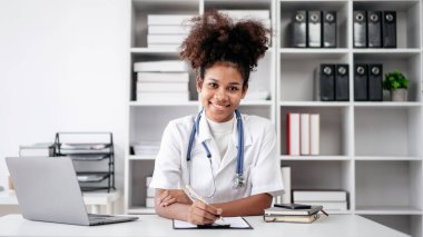 Beyaz laboratuvar önlüklü bir kadın, elinde dizüstü bilgisayar ve not panosuyla bir masada oturuyor. Gülümsüyor ve mutlu.