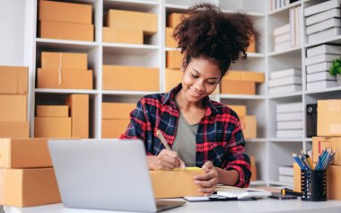 Masasında dizüstü bilgisayarı ve kutuları olan bir kadın oturuyor. Sarı kalemli bir kağıt parçasına yazıyor.