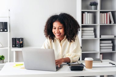 Masasında dizüstü bilgisayarı ve hesap makinesi olan bir kadın oturuyor. Gülümsüyor ve mutlu.