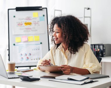 Arkasında beyaz bir tahta olan bir kadın masada oturuyor. Gülümsüyor ve bir panoya yazıyor. Arkasındaki beyaz panoda bir sürü not ve diyagram var.