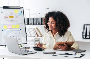 Bir kadın elinde dizüstü bilgisayar ve not panosuyla masada oturuyor. Gülümsüyor ve mutlu.