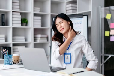 Masasında dizüstü bilgisayarı ve mavi rozeti olan bir kadın oturuyor. Beyaz bir gömlek giyiyor ve acı çekiyor.