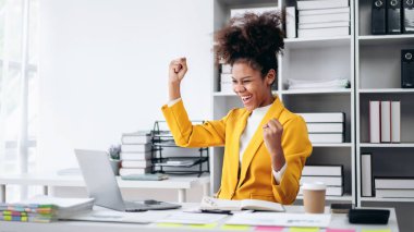 Sarı ceketli bir kadın ellerini havaya kaldırarak kutlama yapıyor. O bir dizüstü bilgisayar ve bir fincan kahveyle bir masada oturuyor.
