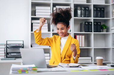 Sarı ceketli bir kadın ellerini havaya kaldırarak kutlama yapıyor. O bir dizüstü bilgisayar ve bir fincan kahveyle bir masada oturuyor. Başarı ve mutluluk kavramı