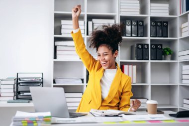 Sarı ceketli bir kadın ellerini havaya kaldırarak kutlama yapıyor. O bir dizüstü bilgisayar ve bir fincan kahveyle bir masada oturuyor. Başarı ve mutluluk kavramı
