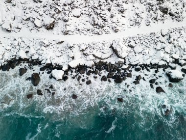 Mavi okyanus dalgalarıyla dolu vahşi bir sahilin yanındaki karda yol. Norveç 'teki tehlikeli kış yolunun havadan görünüşü.